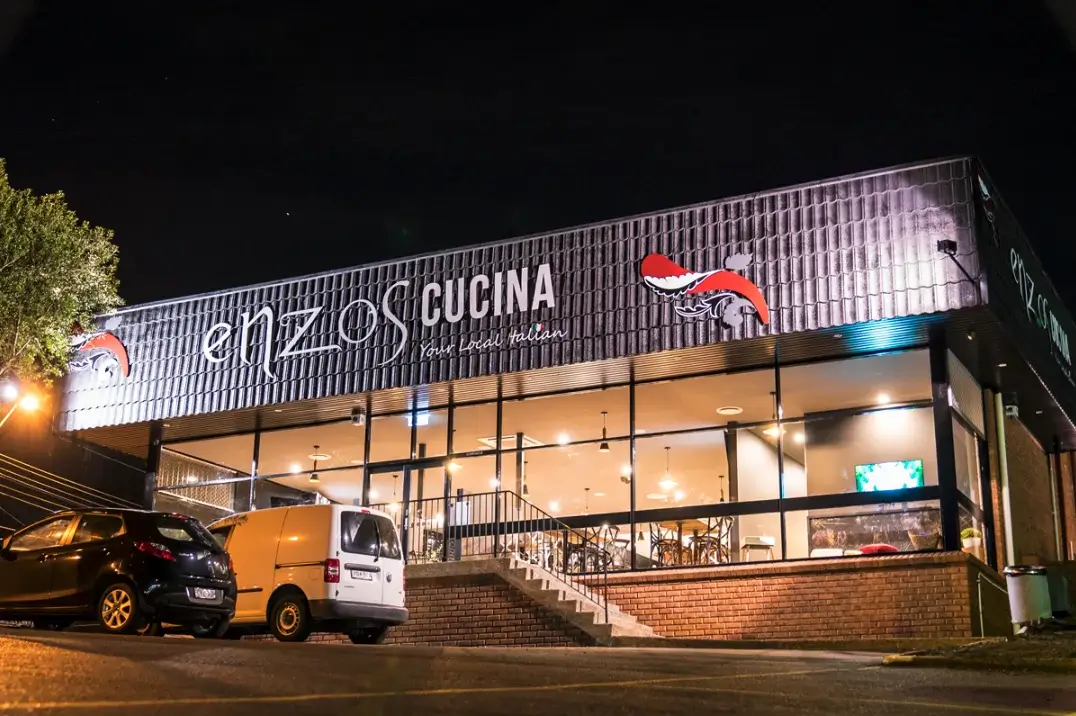 Exterior night view of Enzo’s Cucina restaurant with illuminated signage and parked cars.